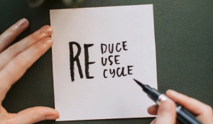 A hand holding a pen over a piece of paper that reads reduce, reuse, recycle
